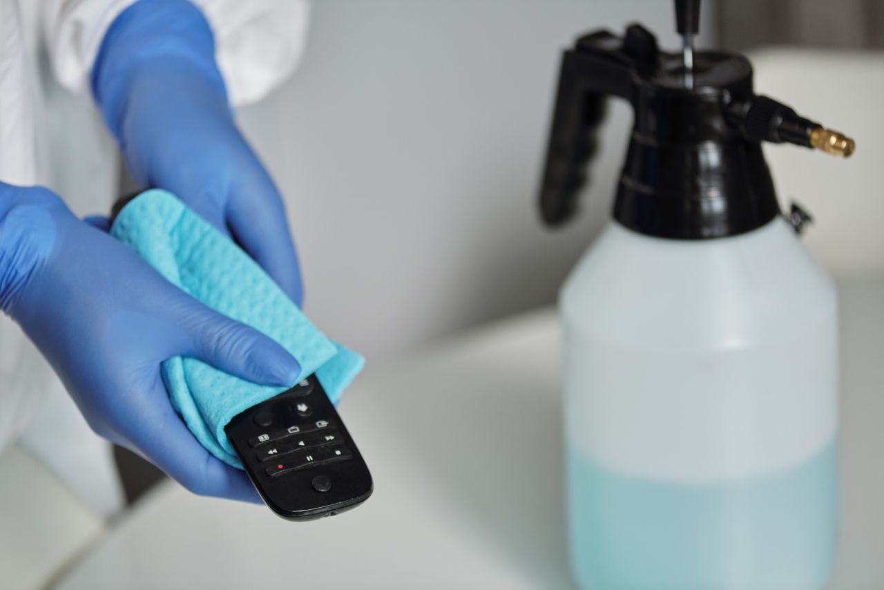 who-we-are Close-up of gloved hands disinfecting a remote control with a cloth and spray bottle.
